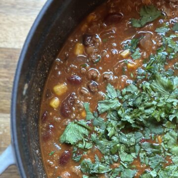 Large Staub pot of homemade chili with cilantro garnishing on wooden kitchen countertop.