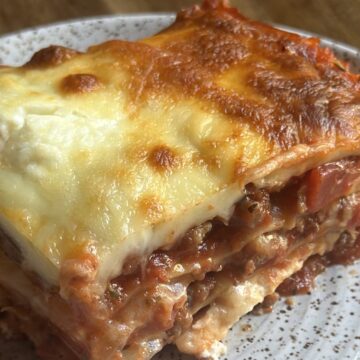 Homemade lasagna slice on white plate served on wooden kitchen counter top.